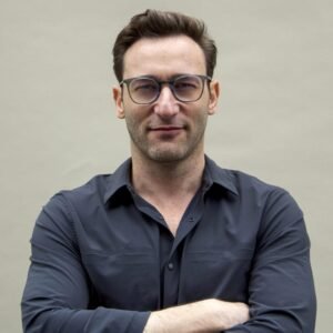 Simon Sinek in a dark button-up shirt and modern glasses, arms crossed, in a professional studio portrait.